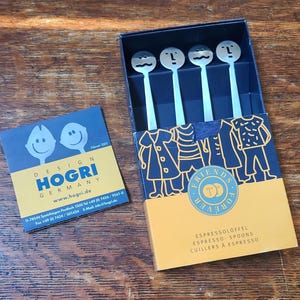 May include: A set of four stainless steel espresso spoons with smiling face designs, presented in a box with a blue and orange design. The box features the text "FRIENDS FOREVER" and illustrations of clothing. A business card with the "HOGRI GERMANY" logo is also visible.