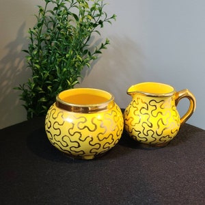 以下が含まれることがあります： 金色の渦巻き模様が施された黄色の砂糖入れとクリーマーのセット。セラミック製で、ビンテージな雰囲気です。