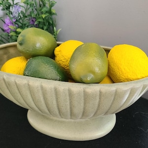 May include: A light green ceramic bowl filled with lemons and limes. The bowl has a pedestal base and a textured, ridged exterior. Green and purple foliage is visible in the background.