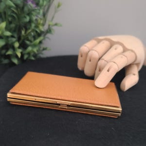 May include: A rectangular, tan leather case with gold-coloured metal trim and a clasp. The case is closed and appears to be empty. The case is on a black surface.