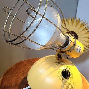 May include: A yellow heat lamp with a protective wire cage around the bulb. The lamp has a rounded base with a dial and warning labels. The lamp is on a wooden surface.