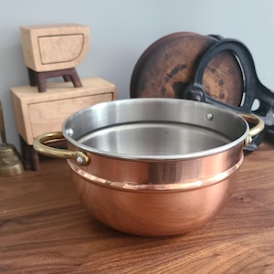 May include: A copper cooking pot with a stainless steel interior and brass handles. The pot is round with a slightly flared rim. The pot is sitting on a wooden surface. Other wooden objects are in the background.