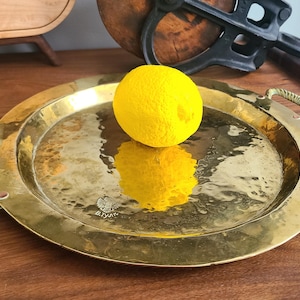 May include: A round, hammered brass tray with handles, holding a yellow sphere. The tray reflects the sphere, creating a mirrored effect. The tray is on a wooden surface.