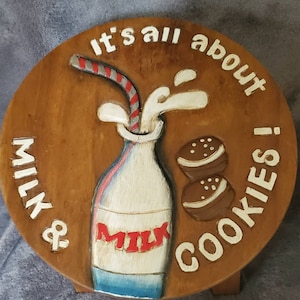 May include: A wooden stool with a round top featuring a carved image of a milk bottle with a straw, milk splashing out, and two chocolate chip cookies. The text "It's all about MILK & COOKIES!" is carved around the image.