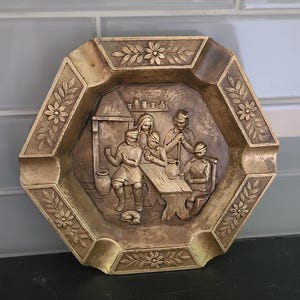 Op de afbeelding: Een vintage, zeshoekige asbak met een gedetailleerde reliëfscène van mensen die rond een tafel verzameld zijn. De asbak heeft een goudkleurige afwerking en bloemen- en bladmotieven langs de randen. De scène toont een familie of een groep mensen.
