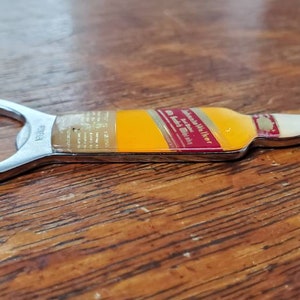 May include: A silver bottle opener shaped like a bottle of Johnnie Walker whisky. The bottle opener has a red and gold label with the text "Johnnie Walker Red Label Blended Scotch Whisky" and "The Authentic Scotch Whisky".