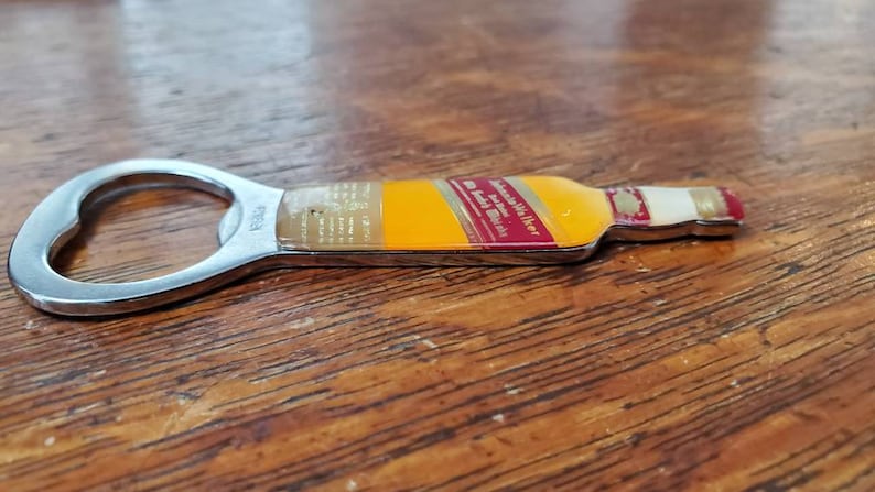May include: A silver bottle opener shaped like a bottle of Johnnie Walker whisky. The bottle opener has a red and gold label with the text "Johnnie Walker Red Label Blended Scotch Whisky" and "The Authentic Scotch Whisky".