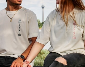 Könnte beinhalten: Ein Paar trägt weiße T-Shirts mit römischen Ziffern auf der Vorderseite. Sie halten sich an den Händen und sitzen auf einer Steinmauer. Der Mann trägt eine goldene Halskette und eine Uhr. Die Frau trägt eine zerrissene Jeans.