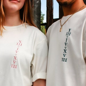 May include: Two white t-shirts with a vertical row of Roman numerals printed on the front. The numerals on the left shirt are in pink, and the numerals on the right shirt are in black.