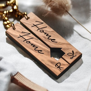 Puede incluir: Un llavero de madera con dos piezas que dicen "Home" y tienen un recorte de una casa. El llavero está unido a una cadena dorada.