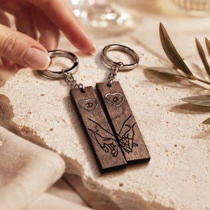 Può includere: Due portachiavi rettangolari in legno scuro con un design inciso al laser di due mani che si tengono. Ogni portachiavi ha un anello e una catena in metallo argentato. Uno mostra la lettera D dentro un cuore, l'altro la lettera C.
