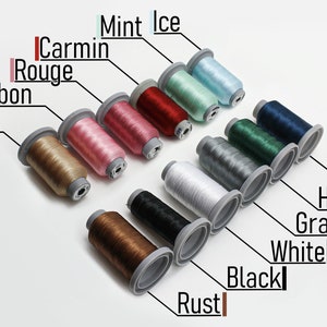 May include: A collection of 12 spools of thread in various colors, including sand, bonbon, carmin rouge, mint ice, rust, black, white, gray, hunter, navy, and two shades of brown. The spools are arranged in a diagonal line from top left to bottom right.