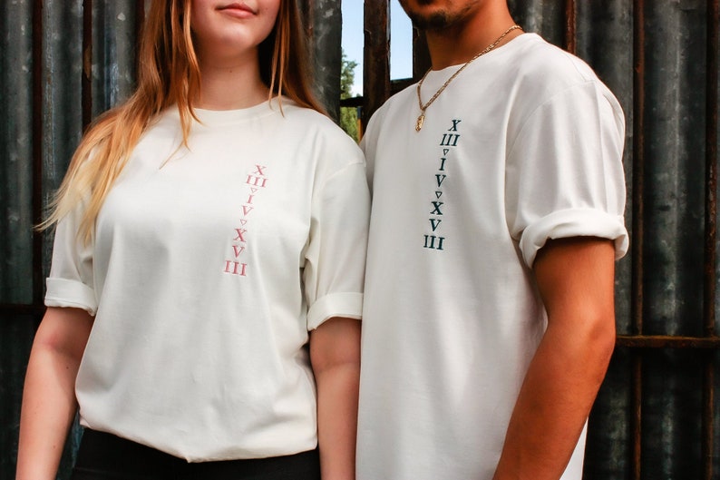 May include: Two white t-shirts with a vertical row of Roman numerals printed on the front. The numerals on the left shirt are in pink, and the numerals on the right shirt are in black.