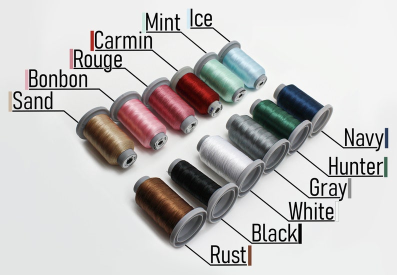 May include: A collection of 12 spools of thread in various colors, including sand, bonbon, carmin rouge, mint ice, rust, black, white, gray, hunter, navy, and two shades of brown. The spools are arranged in a diagonal line from top left to bottom right.