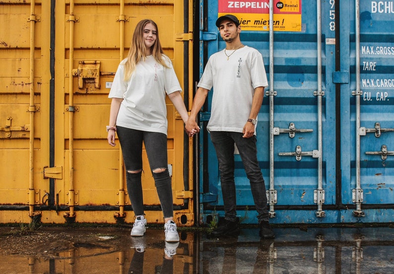 May include: Two people stand in front of a yellow and blue shipping container. They are both wearing white t-shirts and black jeans. The person on the left is holding the hand of the person on the right. The person on the right is wearing a black baseball cap.