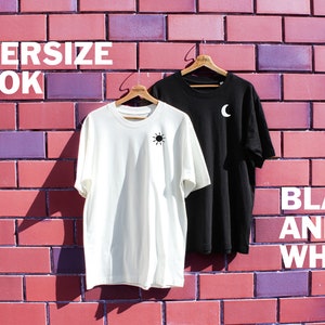 May include: Two oversized t-shirts hanging on wooden hangers against a brick wall. The t-shirt on the left is white with a black sun graphic. The t-shirt on the right is black with a white crescent moon graphic. The text "OVERSIZE LOOK" is at the top of the image and the text "BLACK AND WHITE" is at the bottom of the image.