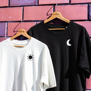 May include: Two t-shirts hanging on wooden hangers against a brick wall. The shirt on the left is white with a black sun graphic. The shirt on the right is black with a white crescent moon graphic. Both shirts have the word "MANTIKA" printed on the hanger.