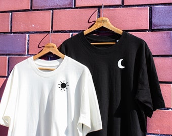 May include: Two t-shirts hanging on wooden hangers against a brick wall. The shirt on the left is white with a black sun graphic. The shirt on the right is black with a white crescent moon graphic. Both shirts have the word "MANTIKA" printed on the hanger.
