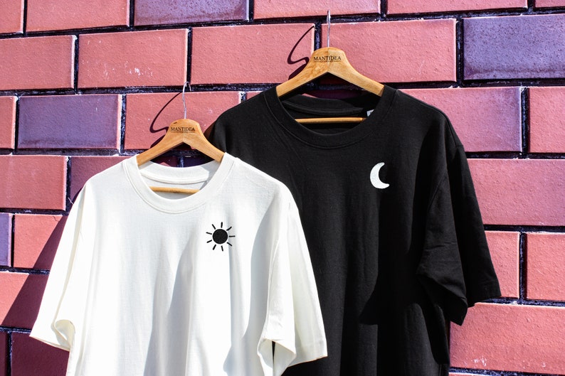May include: Two t-shirts hanging on wooden hangers against a brick wall. The shirt on the left is white with a black sun graphic. The shirt on the right is black with a white crescent moon graphic. Both shirts have the word "MANTIKA" printed on the hanger.