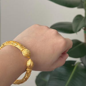 May include: A gold-colored bangle bracelet with intricate floral and scrollwork designs. The bracelet has a hinged opening and decorative finials at each end. The jewelry is displayed against a neutral background with a green plant.