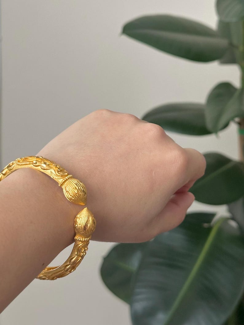 May include: A gold-colored bangle bracelet with intricate floral and scrollwork designs. The bracelet has a hinged opening and decorative finials at each end. The jewelry is displayed against a neutral background with a green plant.