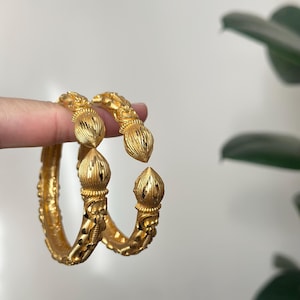 May include: Two gold-colored bangles with intricate floral and textured designs. Each bangle features a decorative, bulbous element at the ends. The jewelry is displayed against a neutral background with a hint of greenery.