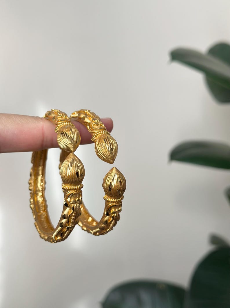May include: Two gold-colored bangles with intricate floral and textured designs. Each bangle features a decorative, bulbous element at the ends. The jewelry is displayed against a neutral background with a hint of greenery.