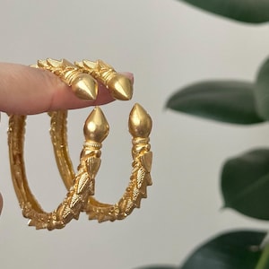 May include: Gold-colored bangles with a textured design and pointed ends. The jewelry has a detailed, ornate pattern and a shiny finish. The bangles are a bright, warm gold color.