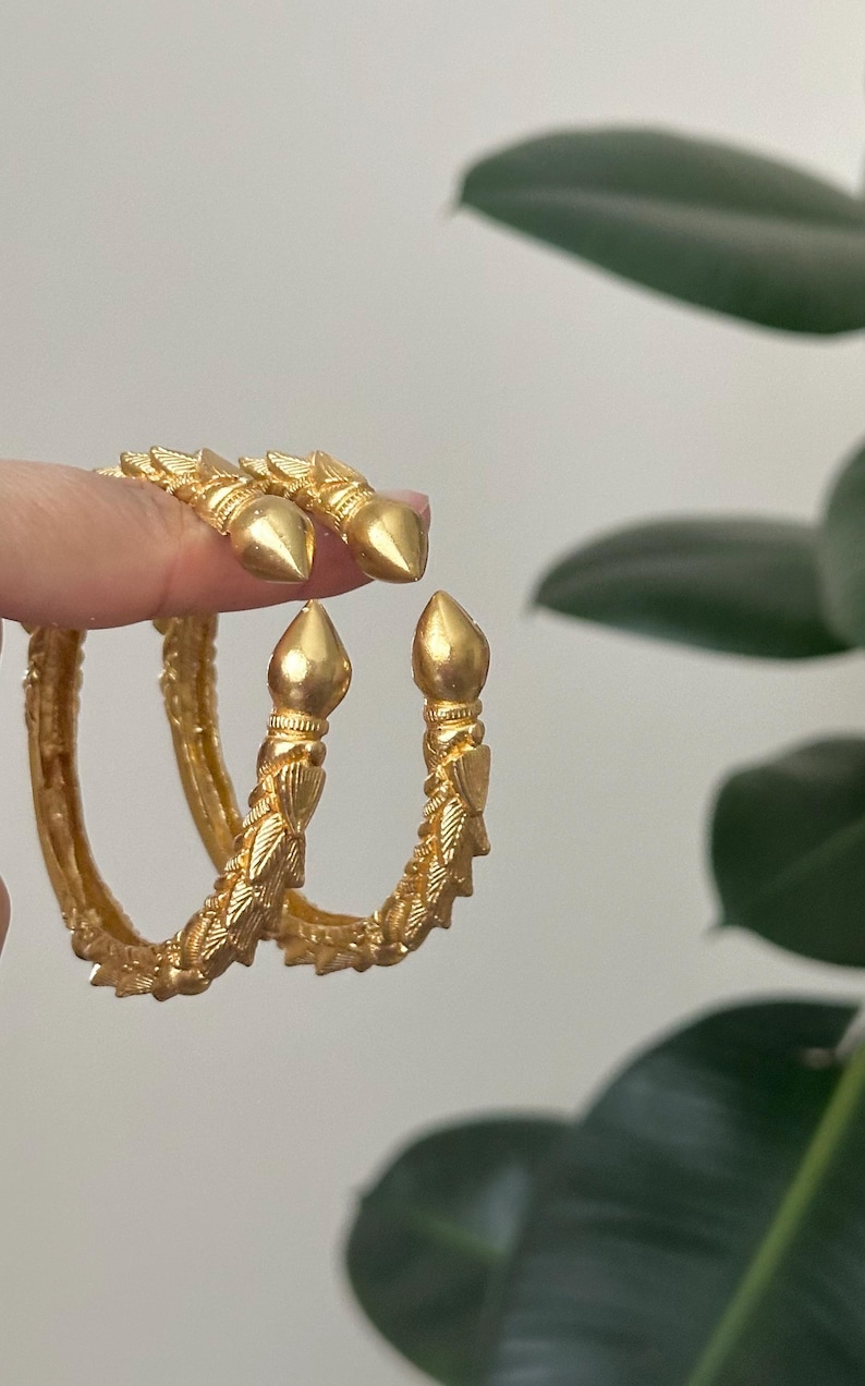 May include: Gold-colored bangles with a textured design and pointed ends. The jewelry has a detailed, ornate pattern and a shiny finish. The bangles are a bright, warm gold color.