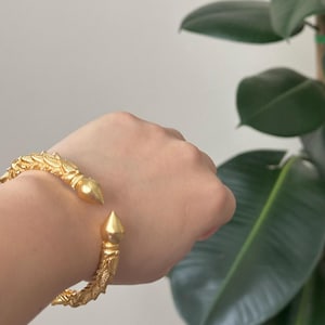May include: Gold-colored cuff bracelet with ornate, textured detailing. The bracelet features a pointed, conical end on each side. The design includes leaf-like patterns and is displayed on a person's wrist, with a green plant in the background.