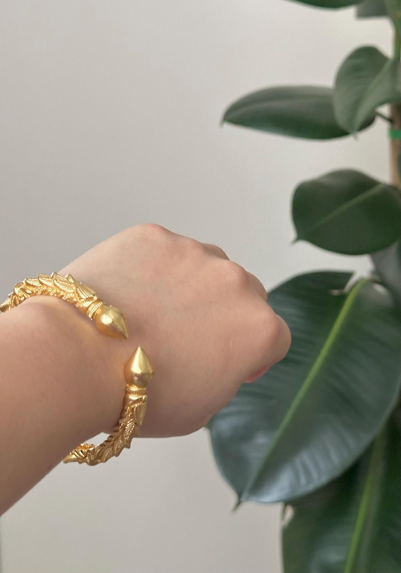 May include: Gold-colored cuff bracelet with ornate, textured detailing. The bracelet features a pointed, conical end on each side. The design includes leaf-like patterns and is displayed on a person's wrist, with a green plant in the background.