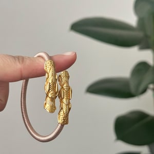 May include: Gold-colored bracelet with detailed, stylized figures at each end. The bracelet has a pinkish-tan, coiled band. The figures appear to be of a mythical or ancient design. The background is a soft, neutral color with green foliage.