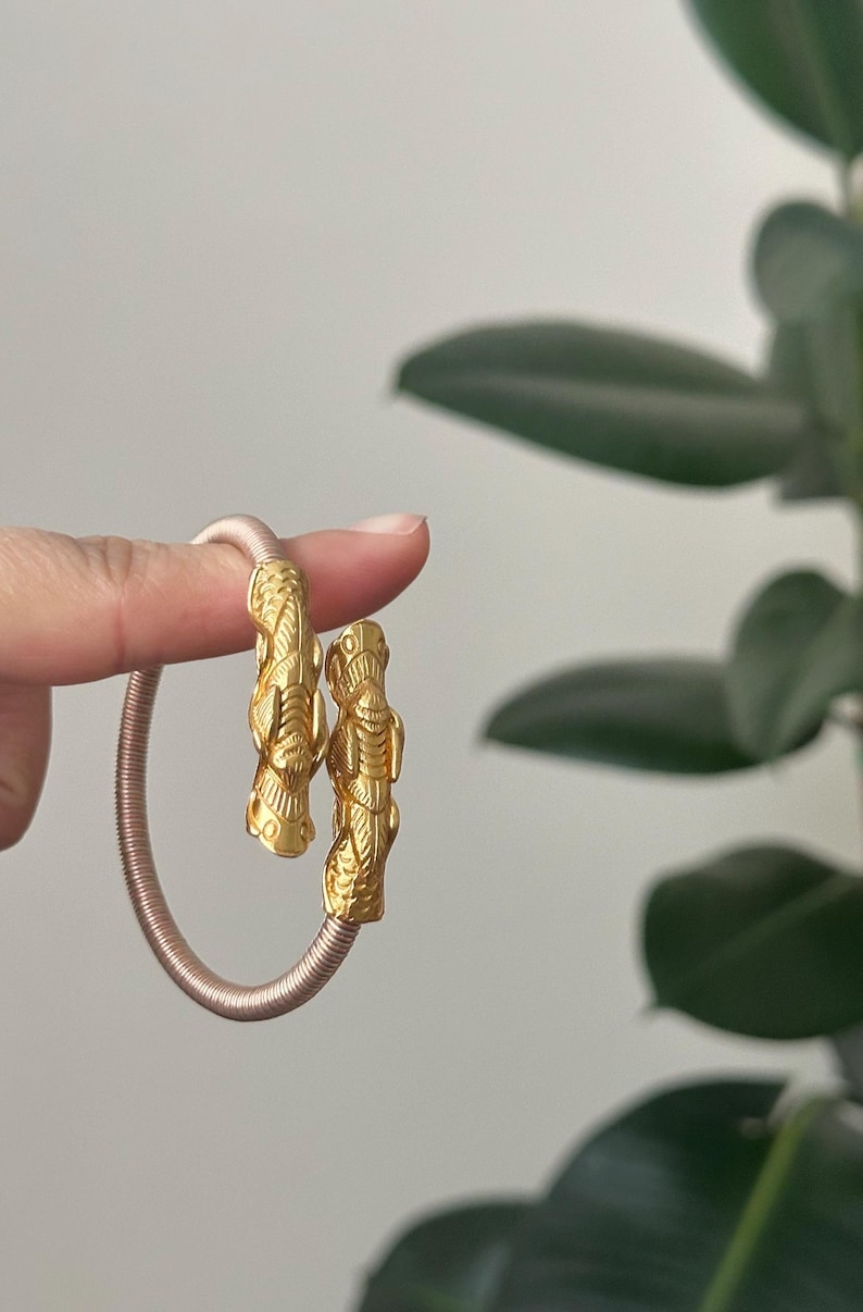 May include: Gold-colored bracelet with detailed, stylized figures at each end. The bracelet has a pinkish-tan, coiled band. The figures appear to be of a mythical or ancient design. The background is a soft, neutral color with green foliage.