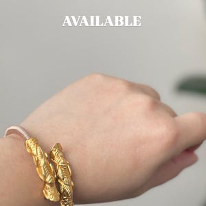 May include: Gold-colored bracelet with intricate, textured design. The bracelet is on a wrist, with the word "AVAILABLE" in white text in the background. The bracelet appears to be made of metal and has a detailed, ornate pattern.