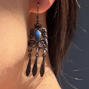 May include: Silver-toned dangle earring with an oval blue stone at the center, surrounded by ornate filigree. Three elongated teardrop-shaped dangles hang below. The earring is worn on the ear.