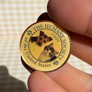 May include: A gold-colored pin from The Humane Society of the United States, featuring two cat portraits and the year 1999. The pin is round with text around the edge.