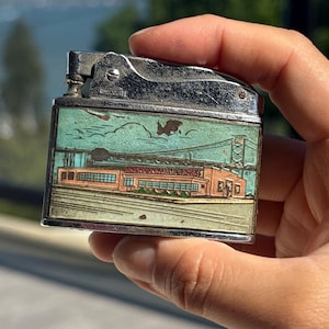 May include: Vintage metal lighter with a detailed illustration of a bridge and buildings in shades of blue, green, and orange. The lighter has a silver-colored top and is held in a hand.