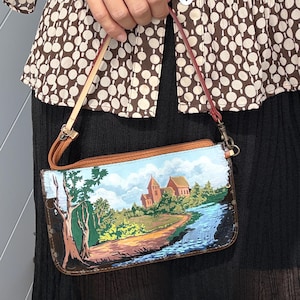 May include: A rectangular wristlet featuring a hand-painted landscape scene of a brick building near a river, surrounded by green trees and a blue sky with clouds. The bag has a brown leather trim, a zipper closure, and a thin, detachable leather strap.