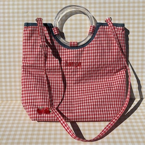 May include: A red and white gingham tote bag featuring a clear circular plastic handle and a long shoulder strap. The word bongo is embroidered in red in the center, and a small red cherry graphic is stitched near the bottom left corner.