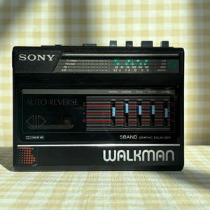 May include: Black Sony Walkman cassette player with the word "WALKMAN" in silver lettering. Features a 5-band graphic equalizer, radio tuner, and "AUTO REVERSE" function. The device is set against a checkered background.