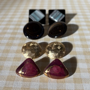 May include: A collection of six pairs of earrings in various shapes and designs. The earrings include square black studs with a marbled pattern, dark brown oval earrings, gold textured ball earrings, and maroon triangle earrings with gold trim.