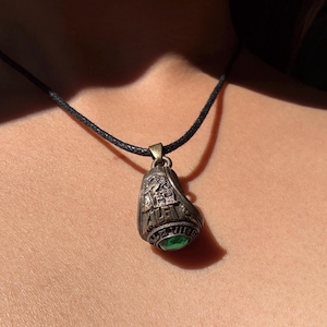 May include: A silver-coloured pendant necklace with a black cord. The pendant is shaped like a ring with the word "Yale" and other text visible. A green gemstone is set in the ring. The necklace is worn around the neck.