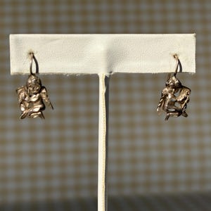May include: A pair of silver-coloured earrings, each with a detailed cherub design. The cherubs have wings and hang from a simple hook. The earrings are displayed on a white stand against a neutral background.