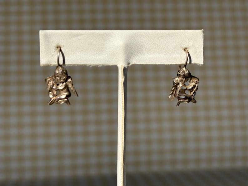May include: A pair of silver-coloured earrings, each with a detailed cherub design. The cherubs have wings and hang from a simple hook. The earrings are displayed on a white stand against a neutral background.
