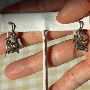 May include: A pair of silver earrings with cherub designs. Each earring shows a seated cherub with wings, playing a musical instrument. The earrings have a hook fastening for pierced ears. The background is a light color.