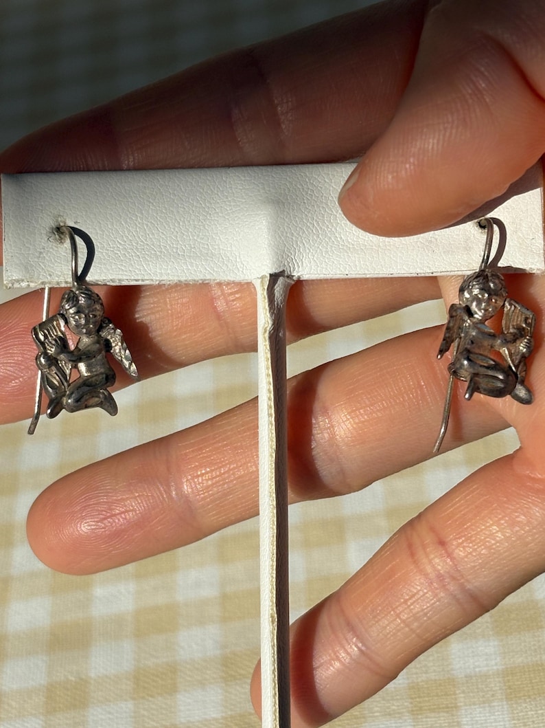 May include: A pair of silver earrings with cherub designs. Each earring shows a seated cherub with wings, playing a musical instrument. The earrings have a hook fastening for pierced ears. The background is a light color.