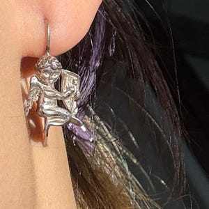 May include: A silver earring depicting a cherub playing a harp. The cherub has detailed wings and is seated on a small chair. The earring is attached to a silver hook. The background includes dark hair and skin.