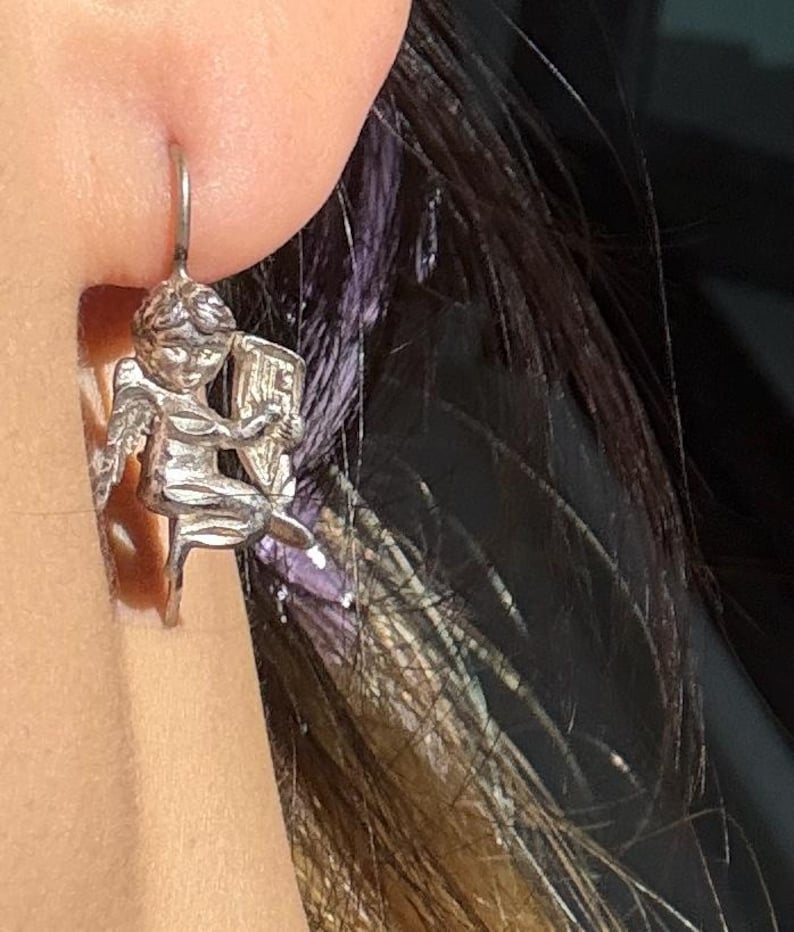 May include: A silver earring depicting a cherub playing a harp. The cherub has detailed wings and is seated on a small chair. The earring is attached to a silver hook. The background includes dark hair and skin.