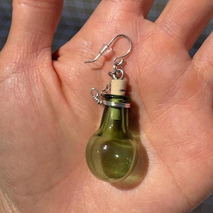 May include: A single green glass bottle earring with a cork stopper and silver-tone metal accents. The earring has a hook closure. The bottle shape is round with a narrow neck.