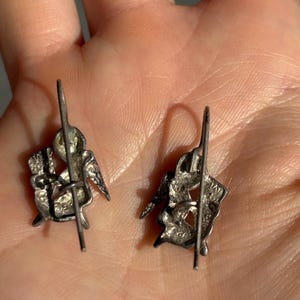 May include: A pair of silver earrings with a detailed design of a seated winged figure, possibly an angel or cherub. The earrings have a post and hook closure, and the design includes intricate detailing.